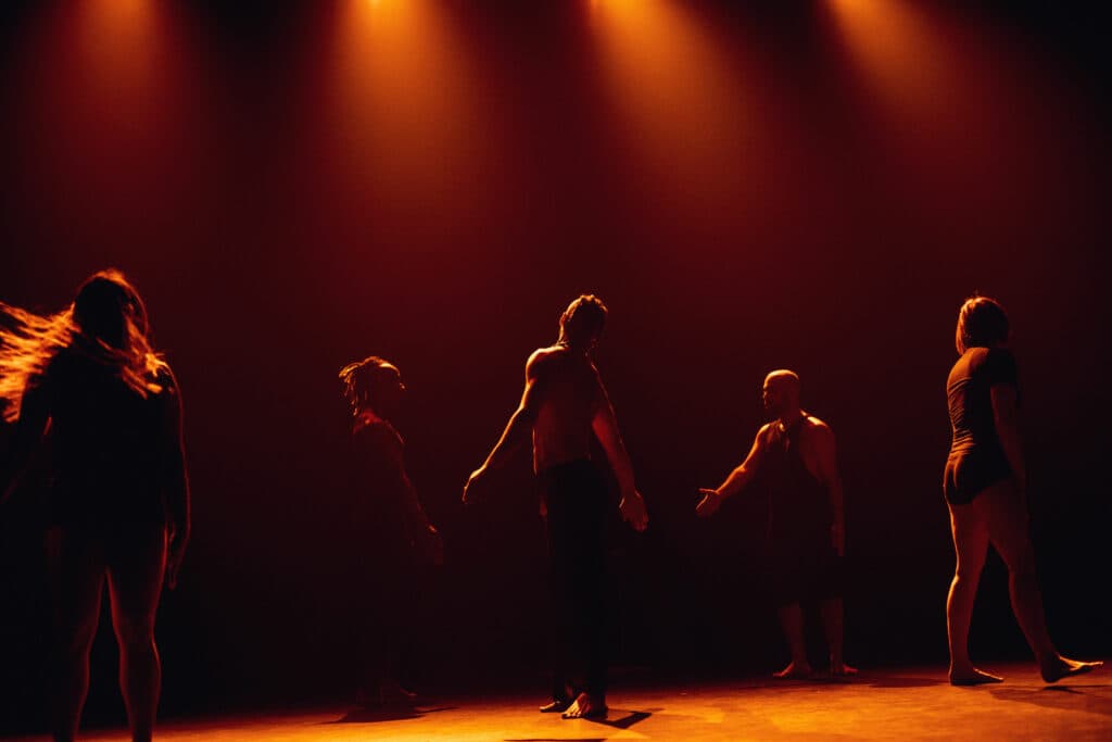 Five performers silhouetted on a dark stage under warm orange spotlights, reaching toward one another