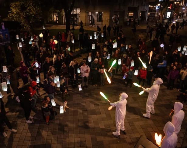 Performers in white hooded suits juggling glowing clubs at night in front of a crowd holding paper lanterns