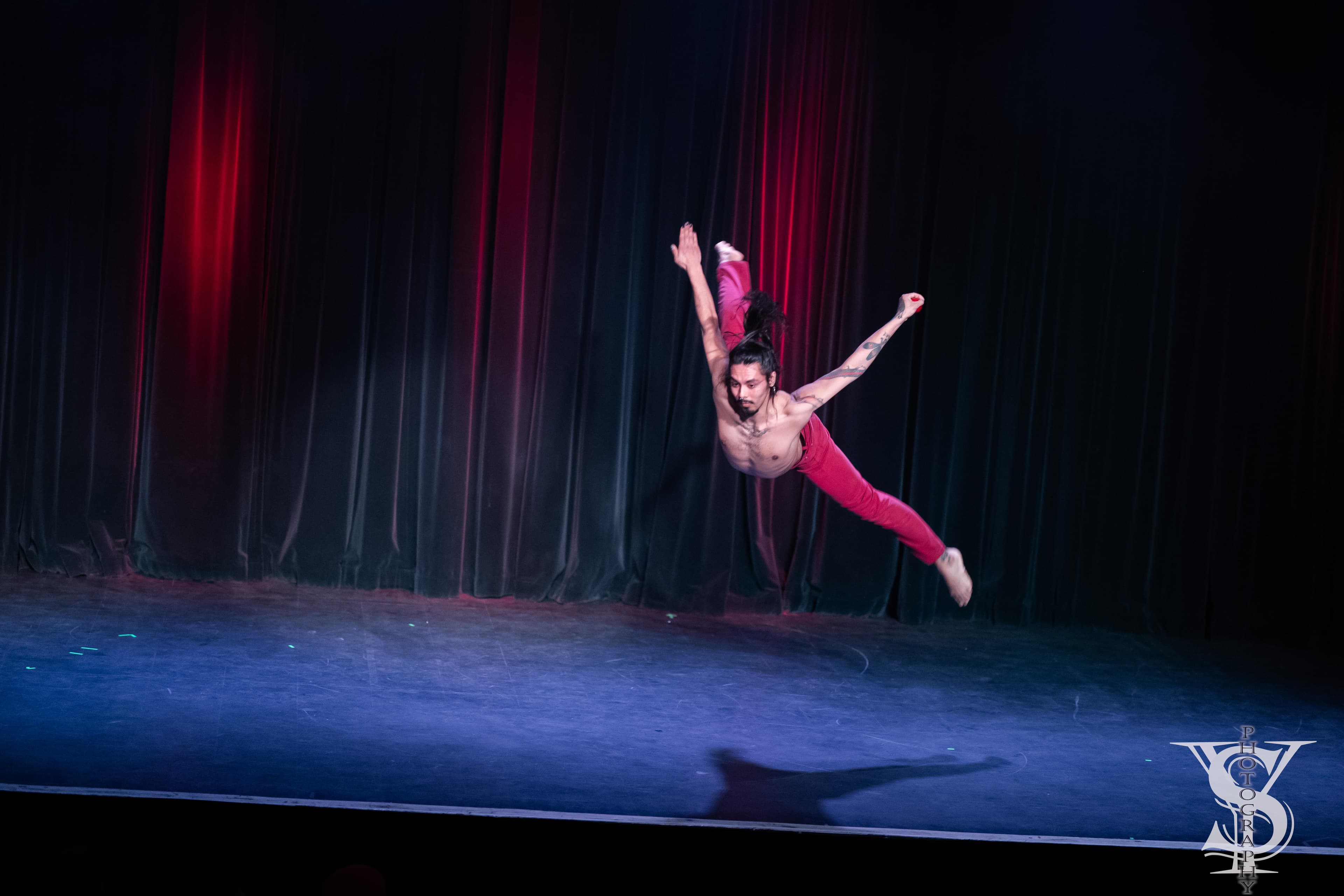 Niki diving horizontally through the air on stage in red trousers, arms outstretched, in front of dark velvet curtains lit red