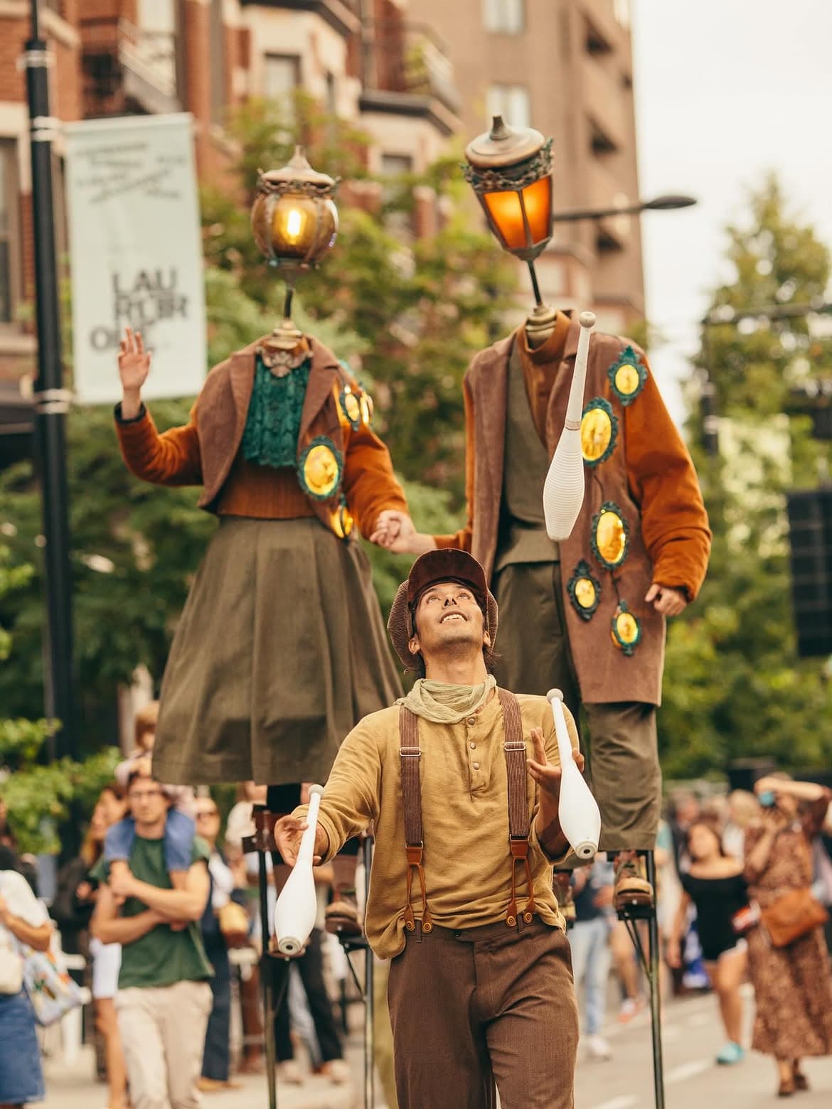 A juggler in a flat cap and suspenders looks up at his white clubs, followed by two stilt performers with glowing lamppost heads