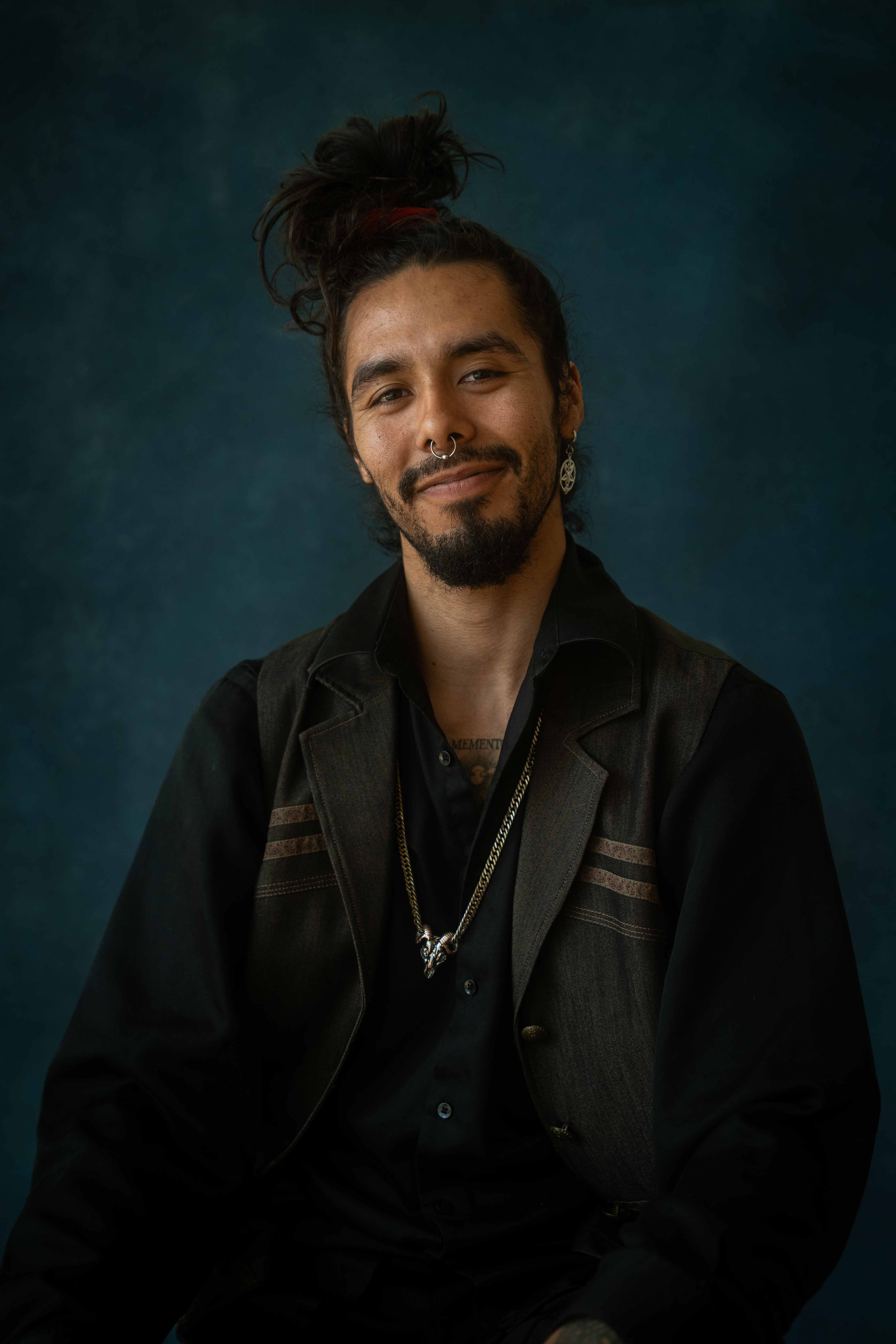Portrait of Niki smiling, hair tied up, wearing a dark vest against a deep teal backdrop